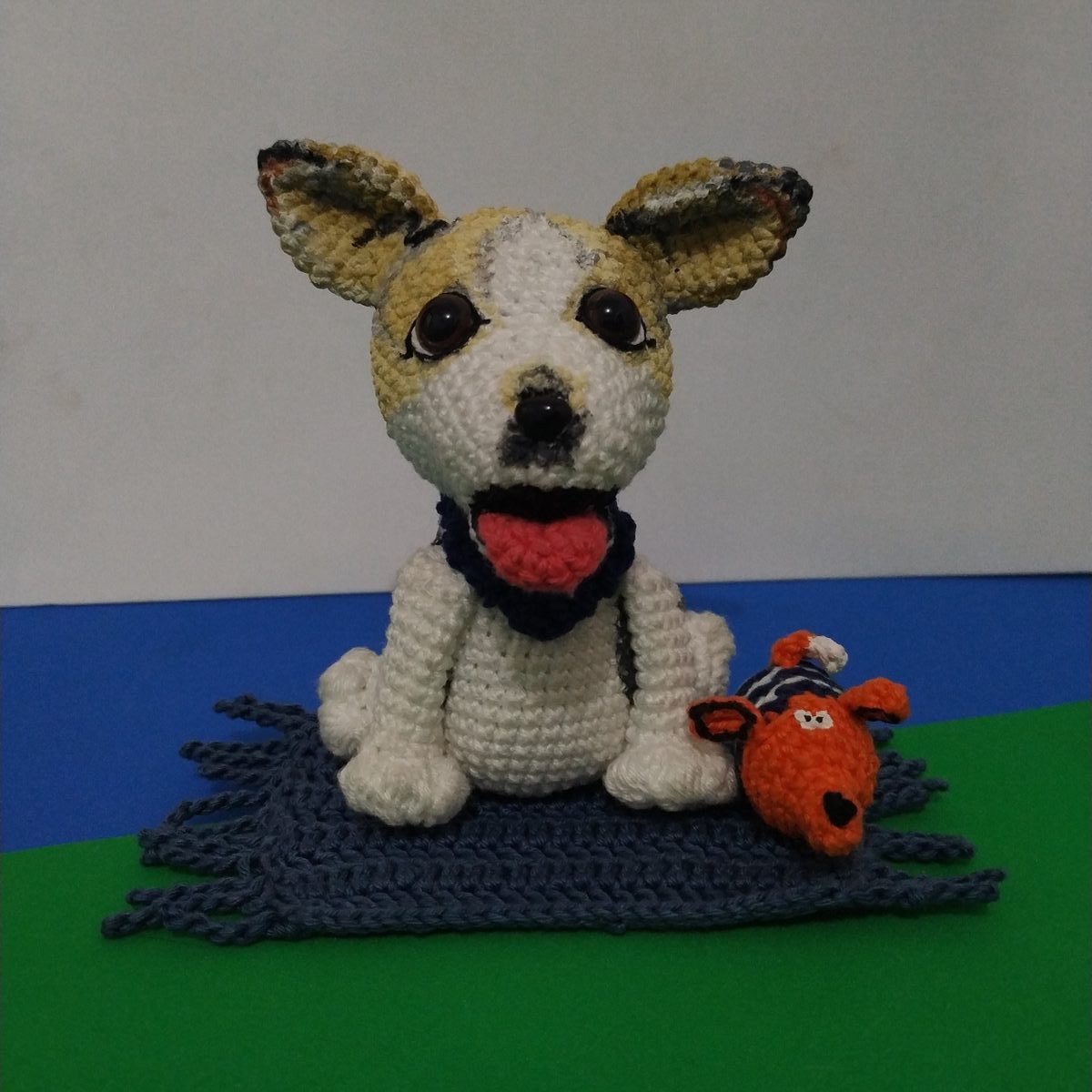 Amigurumi de cachorro em crochê branco e caramelo com bandana azul, ao lado do brinquedo de crochê laranja que era o favorito do pet