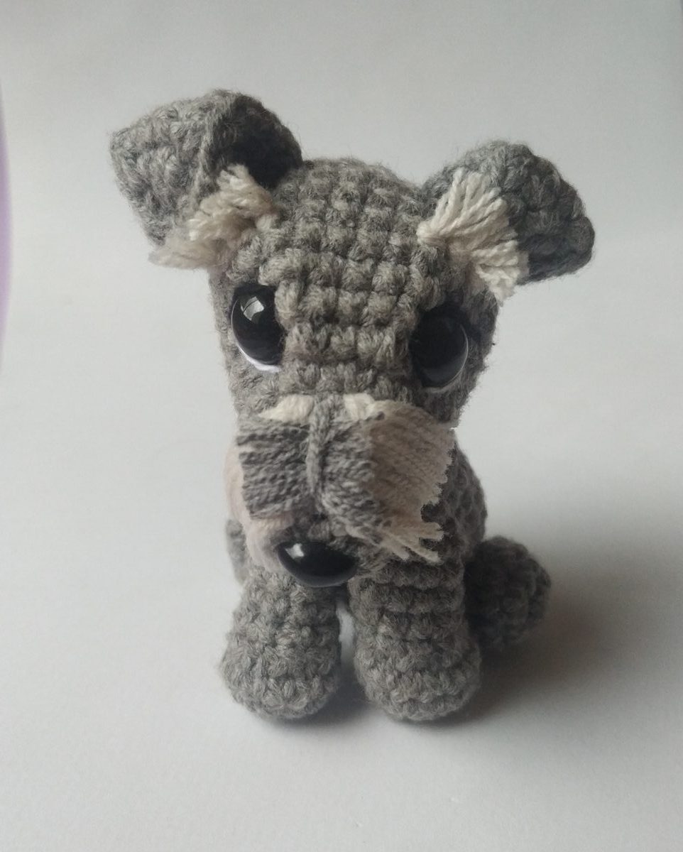 Amigurumi mini de cachorro cinza em crochê com orelhas caídas e franja clara, deitado sobre fundo branco