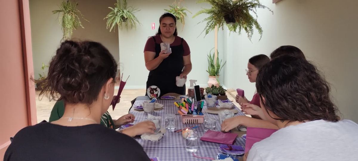 Participantes reunidas ao redor da mesa durante a oficina Cultivo, com Letícia apresentando o plantio de suculentas