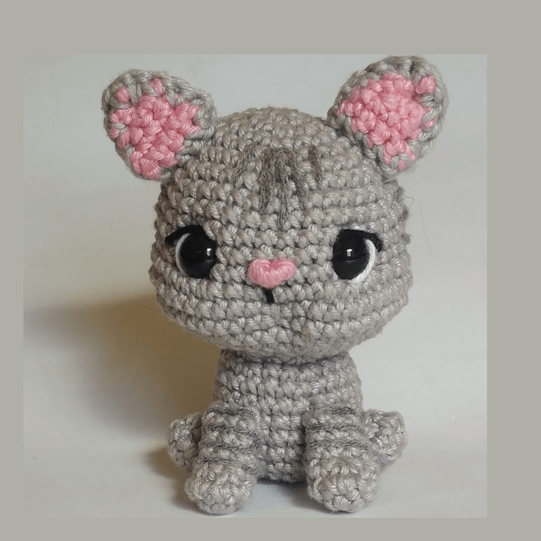 Amigurumi de gato cinza rajado em crochê com orelhas rosa e olhos grandes, sentado sobre fundo claro