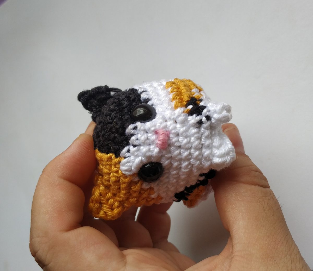 Mão segurando amigurumi mini de gato tricolor branco, laranja e preto feito em crochê