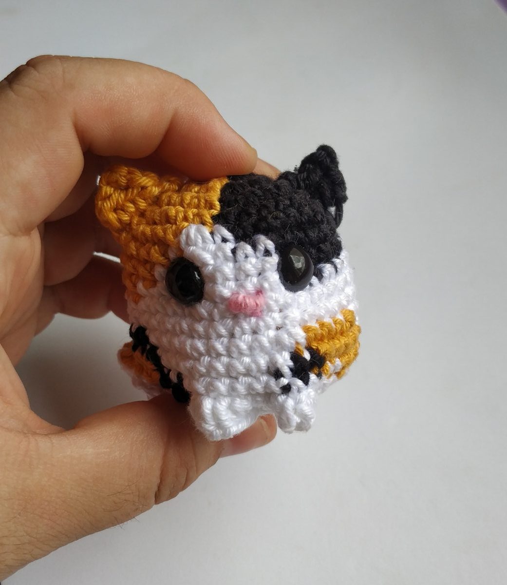 Mão segurando amigurumi mini de gato tricolor branco, laranja e preto feito em crochê