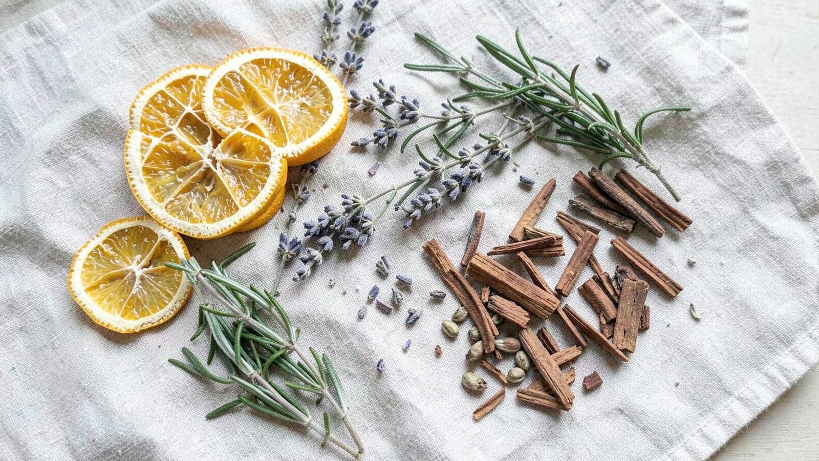 Ingredientes aromáticos naturais sobre pano de linho: rodelas de laranja seca, lavanda, alecrim, lascas de madeira e cardamomo